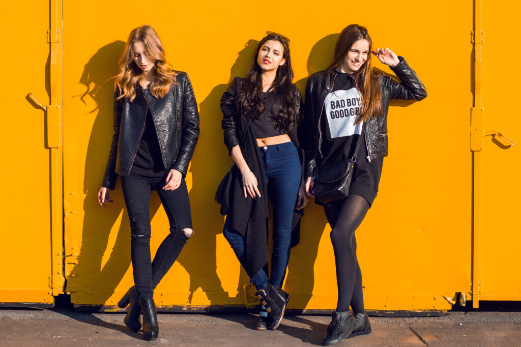 Fashion lifestyle photo of three stylish girls in black spring outfits posing against a pink urban wall.